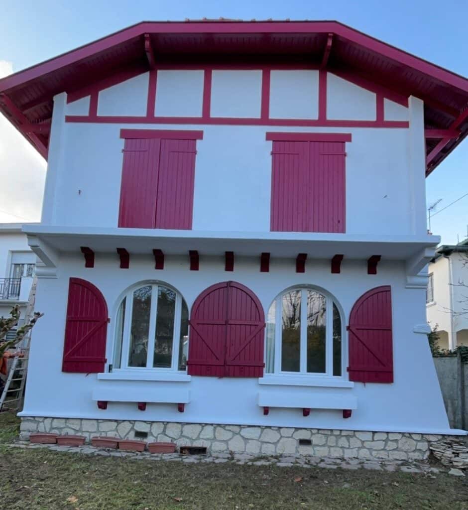 Maison basque rénovée : façade blanche, volets rouges Façade blanche de maison basque rénovée avec boiseries et volets rouges. Fenêtres supérieures à volets fermés, inférieures à arcades ouvertes.