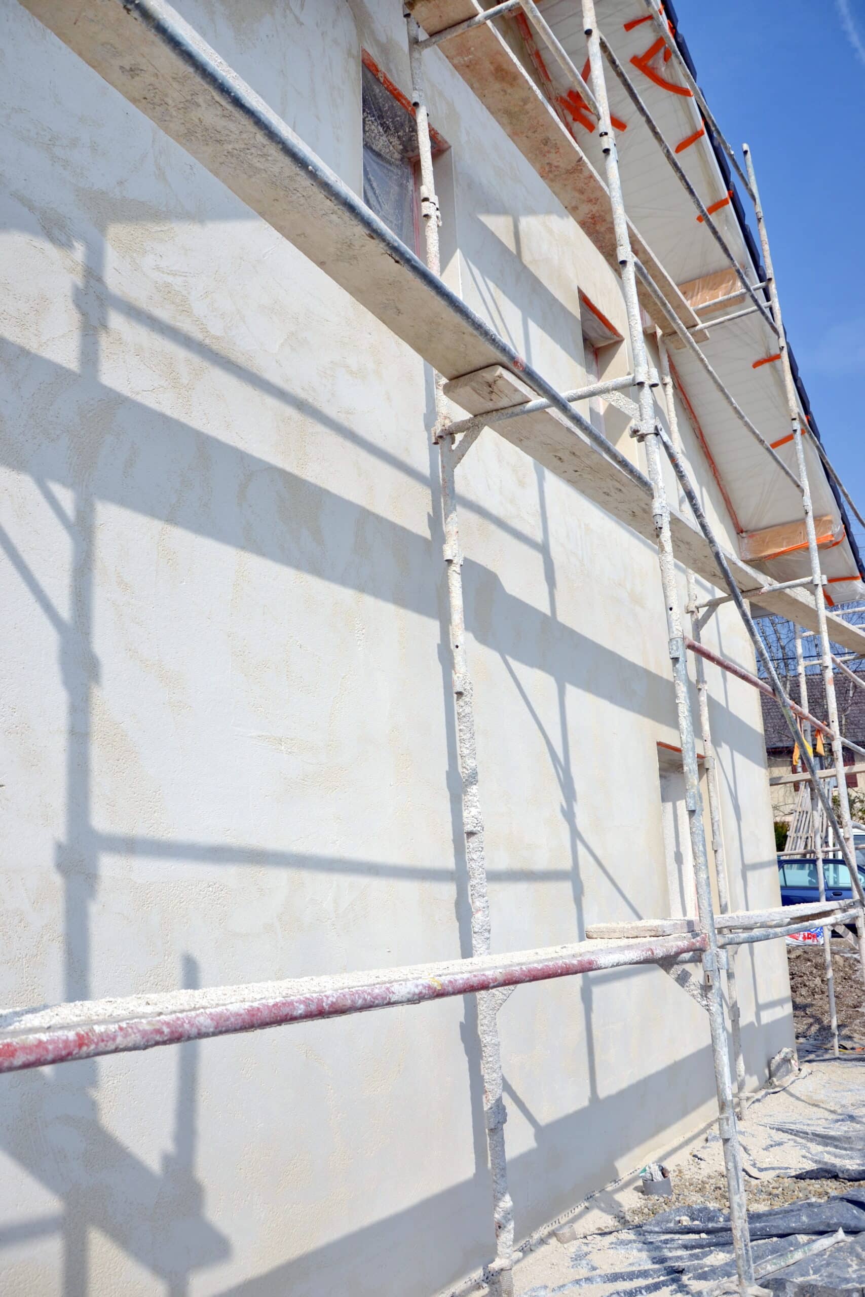 Rénovation façade : échafaudage sur maison enduite Façade de maison en rénovation avec échafaudage et enduit frais. Les ombres de la structure se dessinent sur le crépi clair sous un ciel bleu.