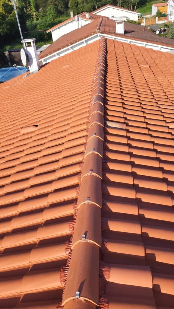 Tuiles Rouges : Faîtage de Toit en Terre Cuite Lumineuse Toit en tuiles de terre cuite rouge, avec faîtage central maintenu par des clips métalliques. Une parabole et des cheminées sont visibles à l'arrière-plan.